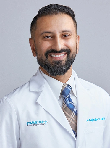 Chiropractor Baljinder Gill smiling in a white coat and tie.
