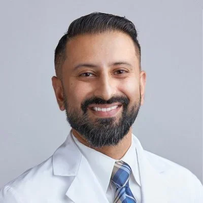 Dr. Baljinder Gill smiling in a white coat and tie.